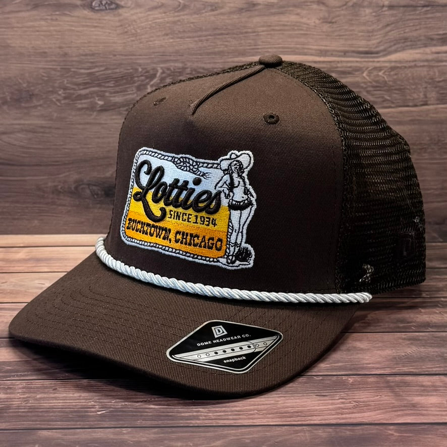 A dark brown trucker hat with black mesh back panels, featuring a front patch that reads 'Clotties Since 1936 Downtown Chicago' with a cowboy illustration, a white rope detail across the brim, and a New Era 9FIFTY sticker on the visor, set against a dark wood background.