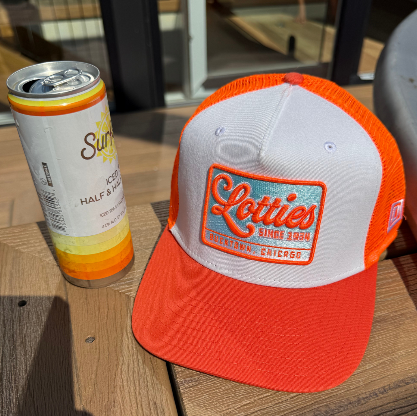 A white and bright orange trucker hat with a 'Scotties Summer Camp' patch, displayed on a wooden deck next to a beverage can.