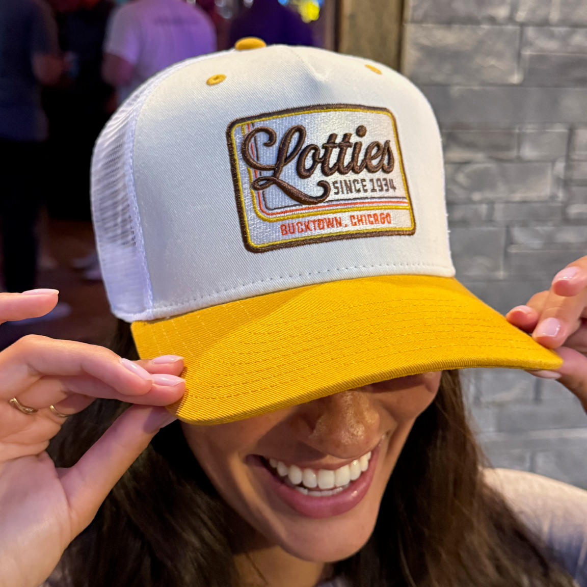 A woman wearing a white and yellow trucker hat with a 'Lotti's Since 1994' patch on the front, holding the brim in front of her eyes in a social setting.