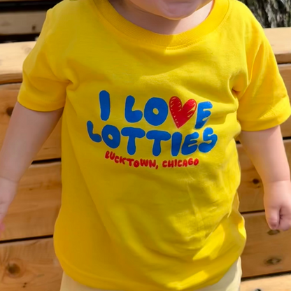 A young toddler girl with her hair in two buns is smiling and wearing a bright yellow t-shirt that reads 'I LOVE LOTTIES' with a red heart and 'BUCKTOWN, CHICAGO' underneath. She is standing outdoors on a wooden bench.