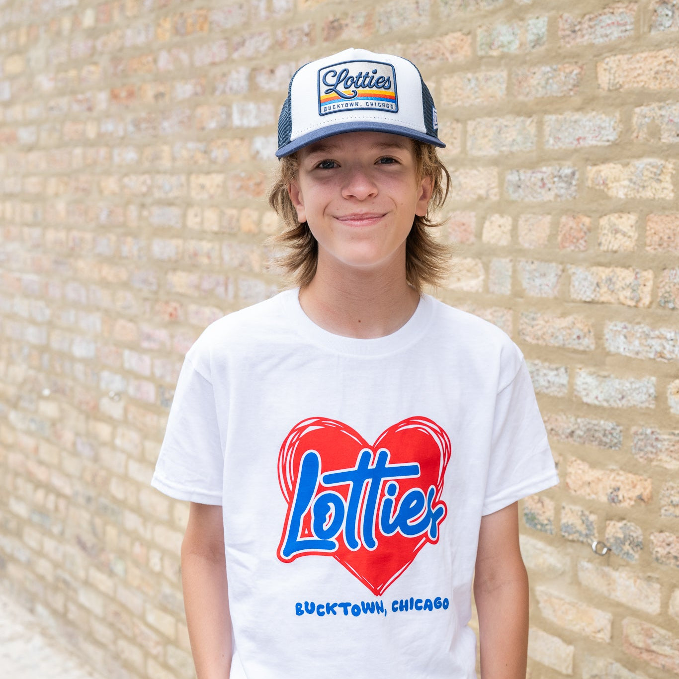 A young man wearing a white crewneck t-shirt with a red and blue 'Lottie Bucktown, Chicago' heart graphic, paired with a blue and white 'Porter' trucker hat, standing against a light brick wall.