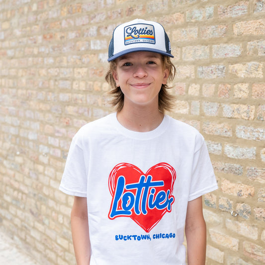 A young man wearing a white crewneck t-shirt with a red and blue 'Lottie Bucktown, Chicago' heart graphic, paired with a blue and white 'Porter' trucker hat, standing against a light brick wall.