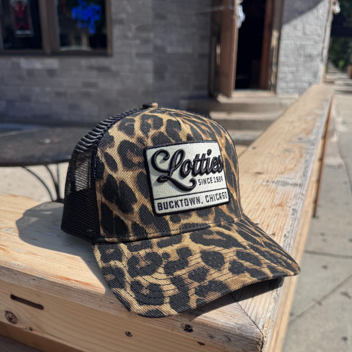 A leopard-print trucker hat with black mesh back panels and a white embroidered patch reading 'Lottie's Backtown, CT' resting on a wooden bench outdoors.