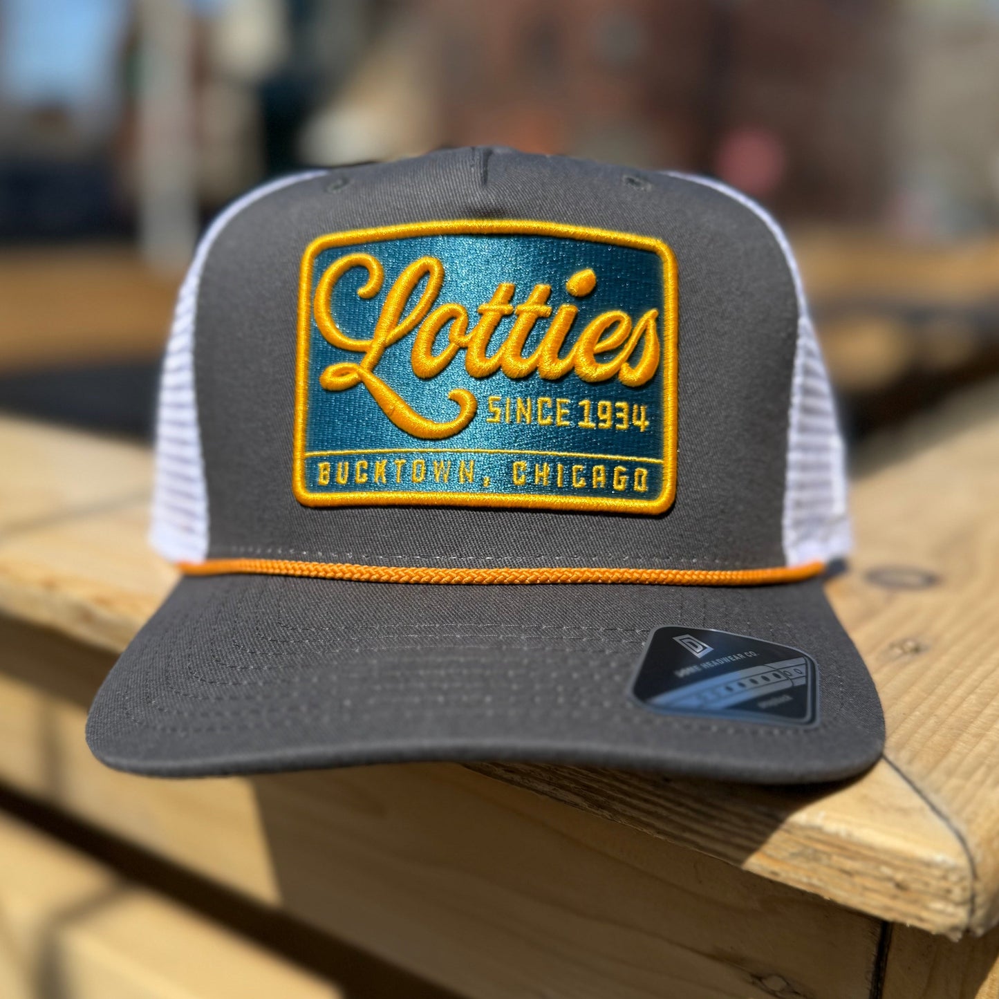 A gray trucker hat with a yellow rope and a signature patch that reads 'Lotties Since 1934' in yellow and teal colors.