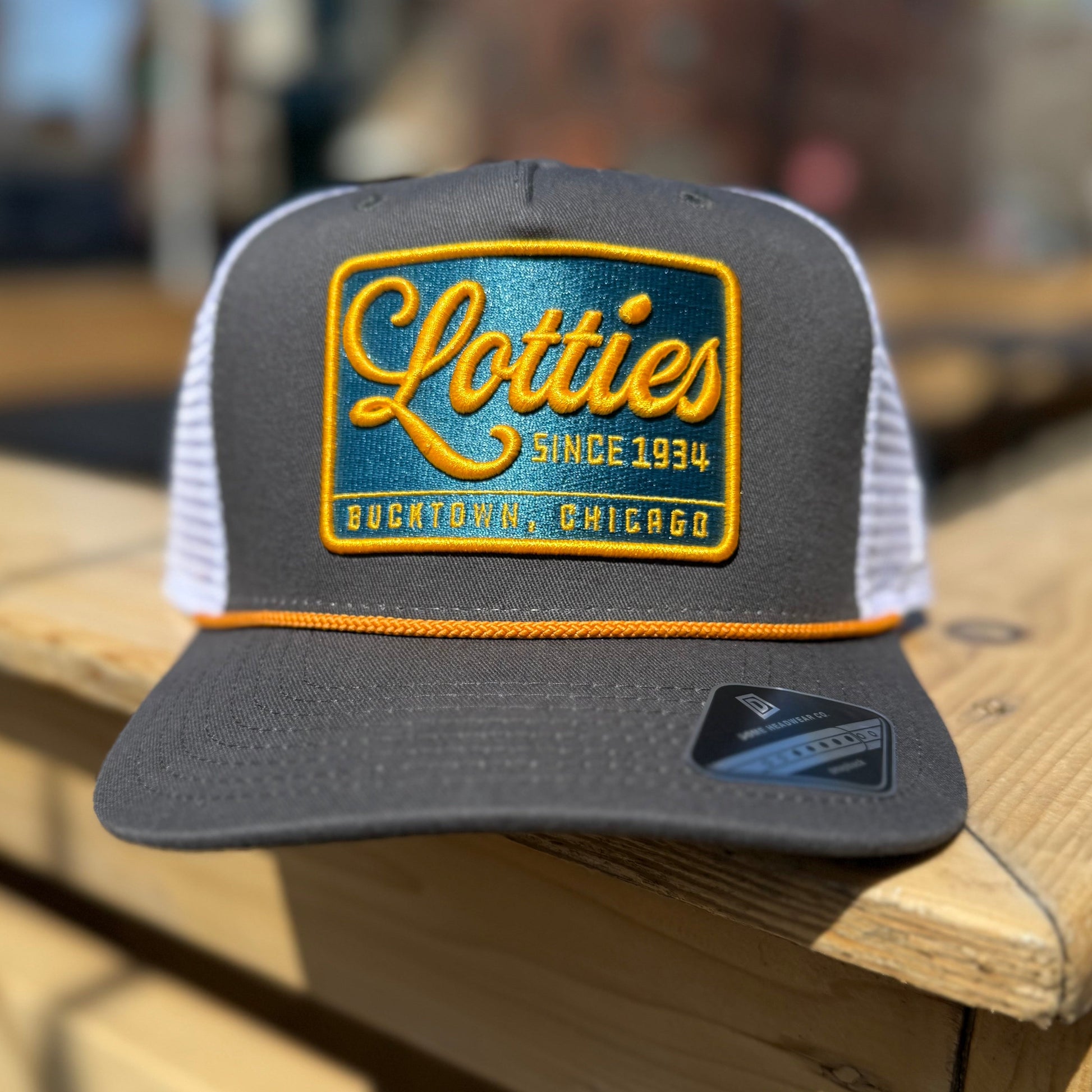 A gray trucker hat with a yellow rope and a signature patch that reads 'Lotties Since 1934' in yellow and teal colors.