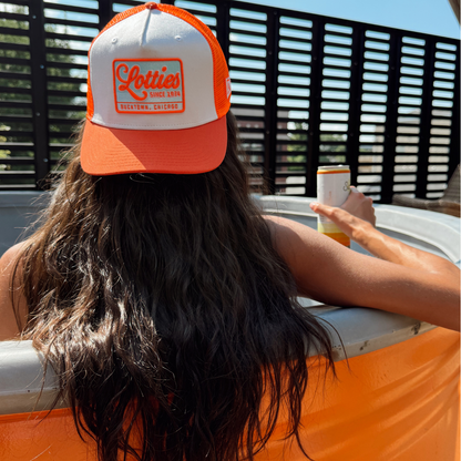 A woman seen from behind wearing an orange and white trucker hat with a 'Letties' logo patch, leaning on an orange cooler outdoors.