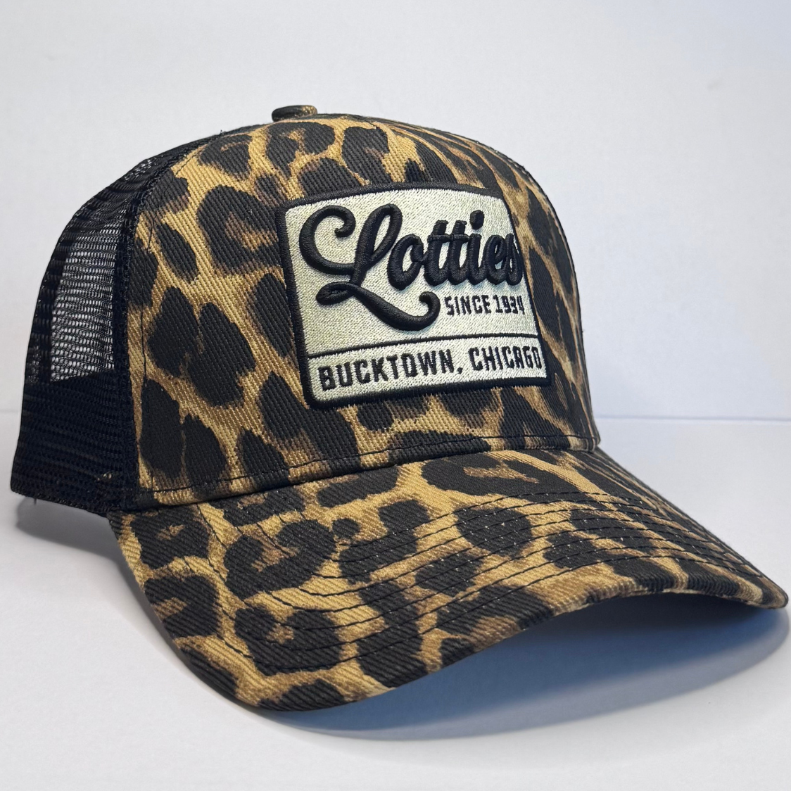 A trucker-style baseball cap with a tan and black leopard print pattern on the front panel and brim, and black mesh back panels. A white rectangular patch on the front reads 'Lotties Since 1981 Bucktown, Chicago.
