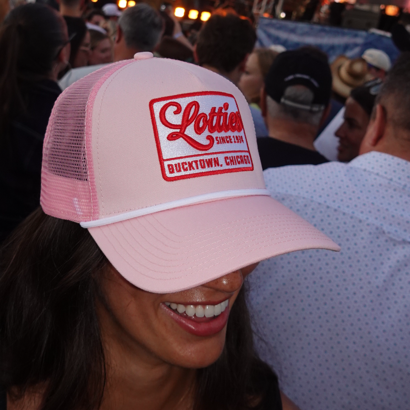 A woman at an outdoor concert wearing a light pink trucker hat with a red and white 'Lethal Injection' logo on the front.